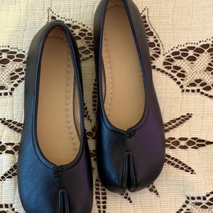 Op de afbeelding: Een paar zwarte split-toe ballerina's met een gladde buitenkant en een beige voering. De schoenen hebben een opvallende verticale inkeping bij de neus, wat zorgt voor een gespleten teenontwerp, en zijn afgewerkt met zichtbare stiksels.
