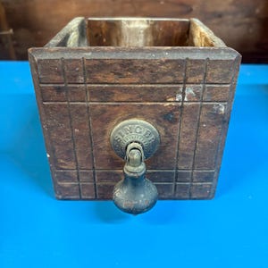 May include: A square, dark brown wooden drawer with a grid-patterned front. It features a central, circular metal plate embossed with the word SINGER and a dark, teardrop-shaped metal pull handle attached to the center of the drawer face.