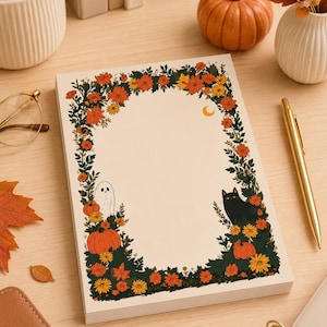 May include: A rectangular notepad featuring a cream-coloured centre framed by a border of dark green foliage, orange and yellow autumn flowers, pumpkins, a small white ghost, a black cat, and a tiny yellow crescent moon in the upper right corner.