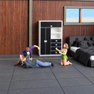 May include: A miniature diorama featuring three small figures in a room. One figure in a purple shirt kneels over another figure in a gray jacket and blue pants. A third figure in a green dress kneels nearby. A white cabinet and a black bed are present.