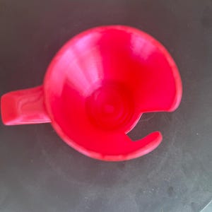 May include: A bright red plastic cup holder featuring a circular base with a side handle and a distinct C-shaped cutout along the rim, designed to accommodate a mug handle while keeping the beverage secure within the circular frame.