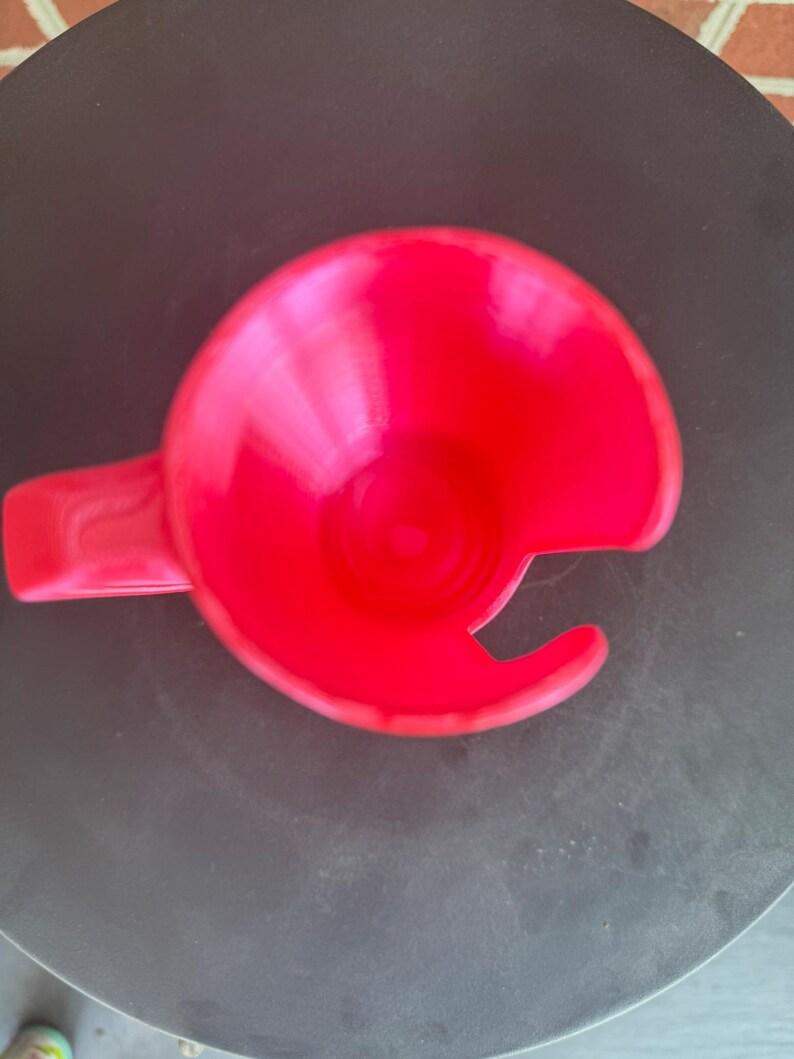 May include: A bright red plastic cup holder featuring a circular base with a side handle and a distinct C-shaped cutout along the rim, designed to accommodate a mug handle while keeping the beverage secure within the circular frame.