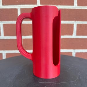 May include: A bright red cylindrical plastic holder featuring a vertical side handle and a long, narrow rectangular cutout running down the length of the body, designed with a smooth, matte finish and an open top and bottom structure.