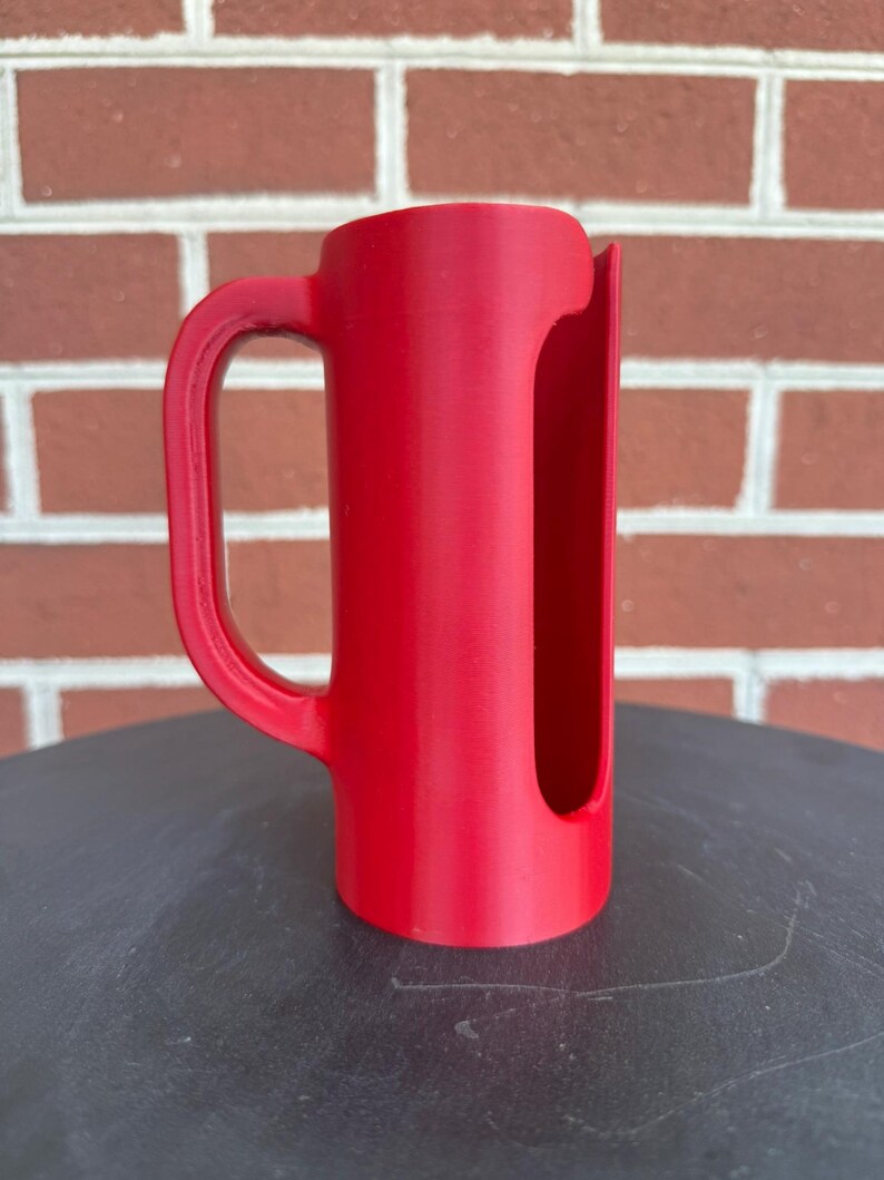 May include: A bright red cylindrical plastic holder featuring a vertical side handle and a long, narrow rectangular cutout running down the length of the body, designed with a smooth, matte finish and an open top and bottom structure.