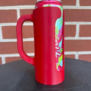May include: A bright red plastic beverage can holder featuring a sturdy side handle. The holder is designed to fit a standard aluminum can, which is partially visible through a vertical cutout, showing a pink, green, and white graphic design.