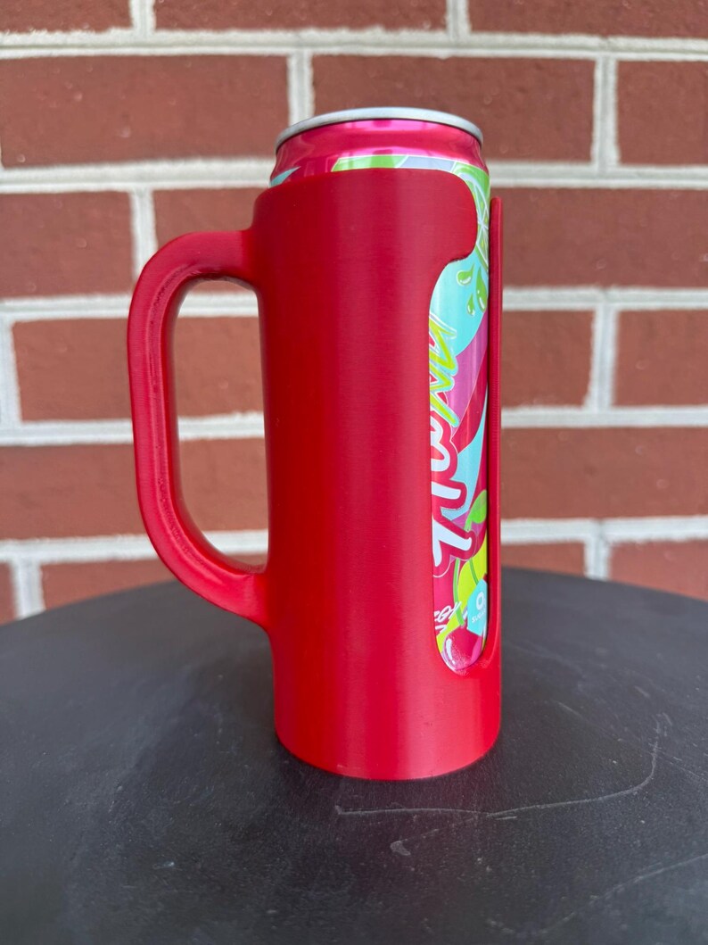May include: A bright red plastic beverage can holder featuring a sturdy side handle. The holder is designed to fit a standard aluminum can, which is partially visible through a vertical cutout, showing a pink, green, and white graphic design.