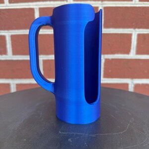 May include: A bright blue cylindrical object with a vertical cutout along one side and a curved handle attached to the exterior. The item features a smooth, solid-colored finish and a hollow interior, with a flat base and a rounded top edge.
