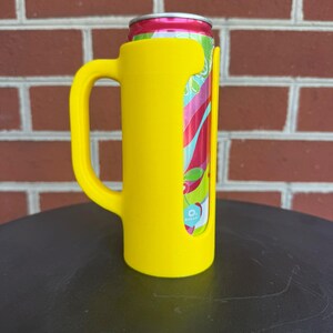 May include: A bright yellow beverage can holder featuring a sturdy side handle. The holder is designed with a vertical cutout that reveals a portion of a colorful aluminum can with red, green, and white graphics and a small blue 0% sugar label.