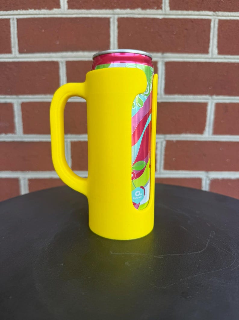May include: A bright yellow beverage can holder featuring a sturdy side handle. The holder is designed with a vertical cutout that reveals a portion of a colorful aluminum can with red, green, and white graphics and a small blue 0% sugar label.