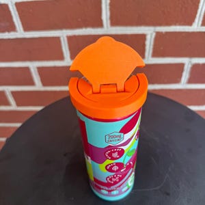 May include: A cylindrical beverage container with a bright orange flip-top lid. The exterior features a light blue, red, and white graphic pattern with text including 200mg CAFFEINE, GLUTEN-FREE, VEGAN, and a cherry illustration on the side.