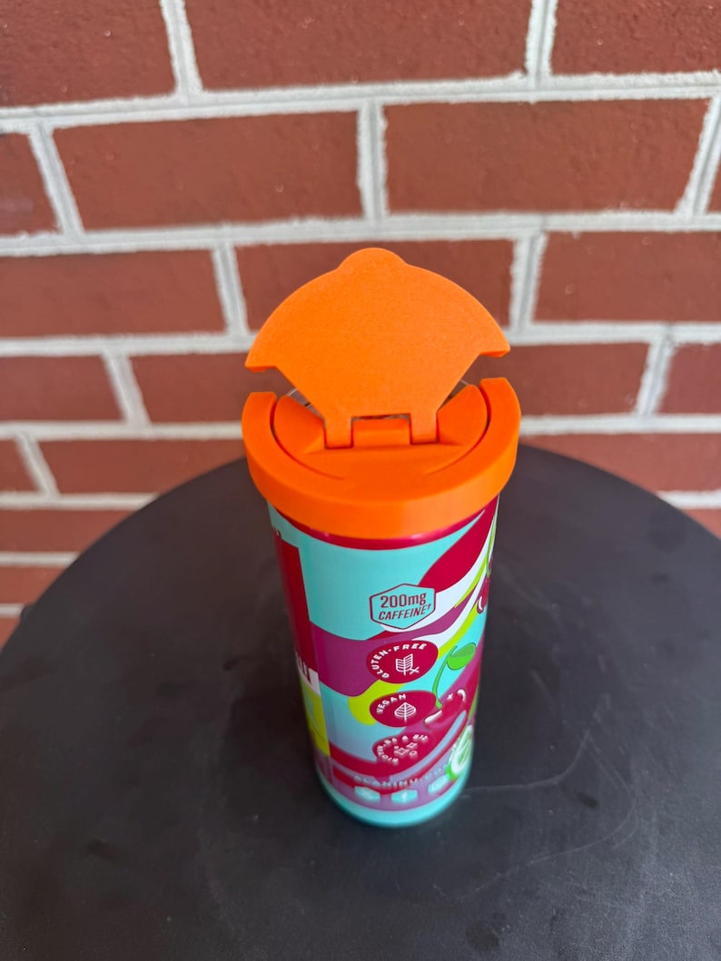 May include: A cylindrical beverage container with a bright orange flip-top lid. The exterior features a light blue, red, and white graphic pattern with text including 200mg CAFFEINE, GLUTEN-FREE, VEGAN, and a cherry illustration on the side.