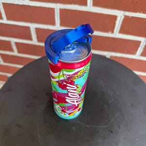 May include: A colorful Alani Nu Cherry Twist energy drink can with a bright red, white, and teal graphic pattern featuring cherry and lime illustrations. A blue plastic lid attachment is secured to the top of the aluminum can lid.