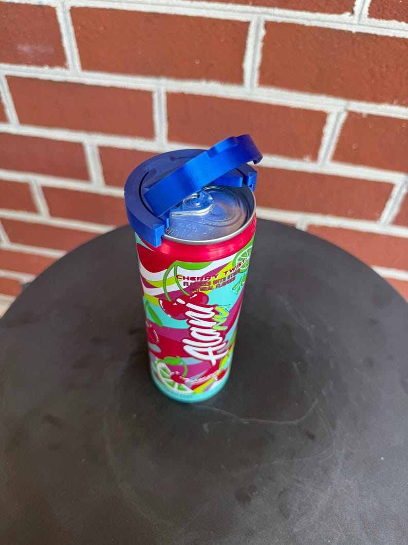 May include: A colorful Alani Nu Cherry Twist energy drink can with a bright red, white, and teal graphic pattern featuring cherry and lime illustrations. A blue plastic lid attachment is secured to the top of the aluminum can lid.