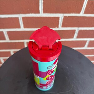 May include: A cylindrical beverage container with a bright red flip-top lid. The body features a light blue background with red and pink accents, displaying text that includes 200mg CAFFEINE, along with circular icons and cherry illustrations.