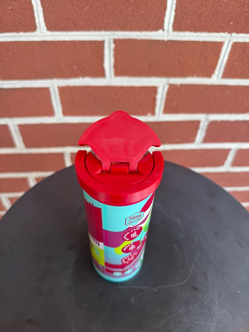 May include: A cylindrical beverage container with a bright red flip-top lid. The body features a light blue background with red and pink accents, displaying text that includes 200mg CAFFEINE, along with circular icons and cherry illustrations.