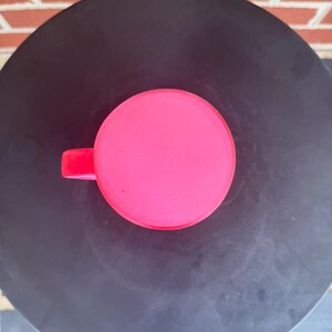May include: A bright neon pink mug viewed from directly above, showing a perfectly circular rim and a small portion of the handle extending to the left side. The mug has a smooth, solid-colored surface and a simple, clean, and functional design.
