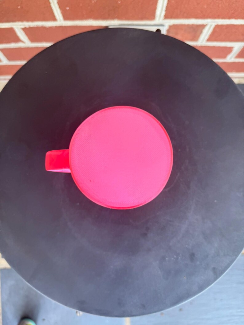 May include: A bright neon pink mug viewed from directly above, showing a perfectly circular rim and a small portion of the handle extending to the left side. The mug has a smooth, solid-colored surface and a simple, clean, and functional design.
