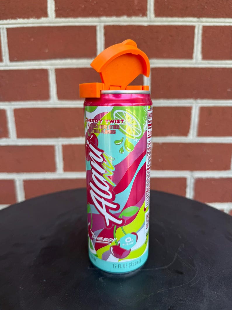 May include: A 12 fl oz aluminum energy drink can labeled Alani Nu Cherry Twist with a bright orange plastic flip-top lid attached to the top. The can features a colorful graphic design with red cherries, lime slices, and green accents on a pink background.