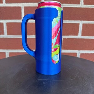 May include: A blue plastic beverage can holder with a side handle, designed to securely fit a slim aluminum can. The can features a vibrant, multi-colored graphic pattern with shades of pink, green, and light blue, and a silver top rim.