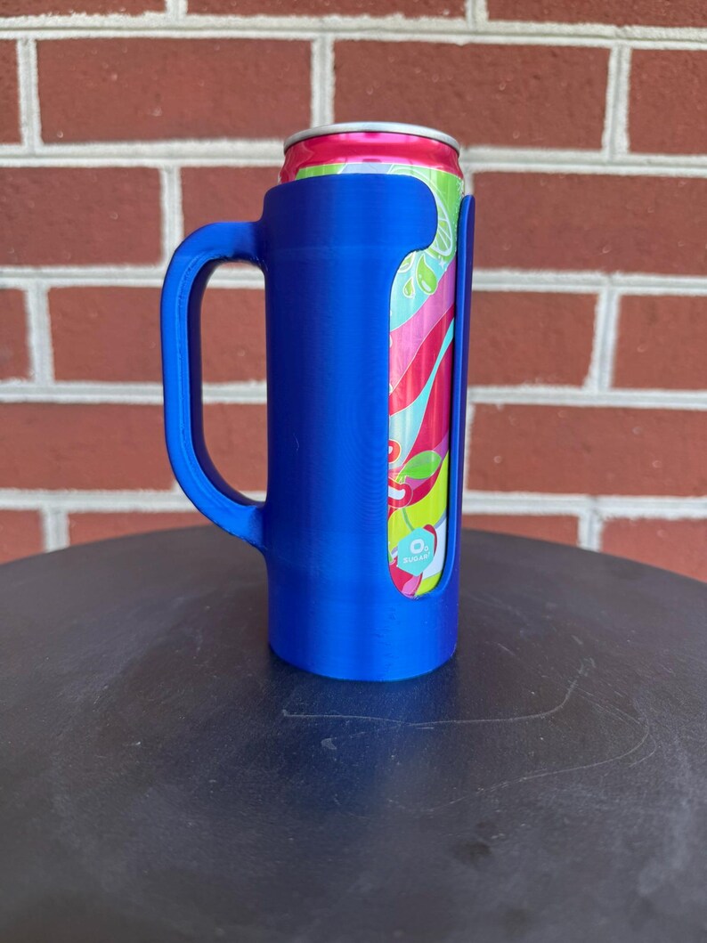 May include: A blue plastic beverage can holder with a side handle, designed to securely fit a slim aluminum can. The can features a vibrant, multi-colored graphic pattern with shades of pink, green, and light blue, and a silver top rim.
