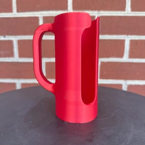 May include: A bright red cylindrical handle attachment featuring a vertical open slot along the side. The object has a curved grip extending from the main body, designed to fit around a container, with a smooth matte finish and a uniform color.