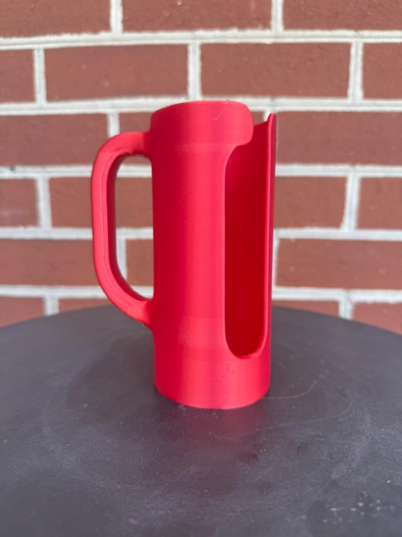 May include: A bright red cylindrical handle attachment featuring a vertical open slot along the side. The object has a curved grip extending from the main body, designed to fit around a container, with a smooth matte finish and a uniform color.