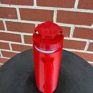 May include: A bright red insulated travel mug with a matching red handle and a hinged lid. The mug features a visible teal-colored inner rim and a cylindrical body design, providing a functional vessel for holding hot or cold beverages while on the go.