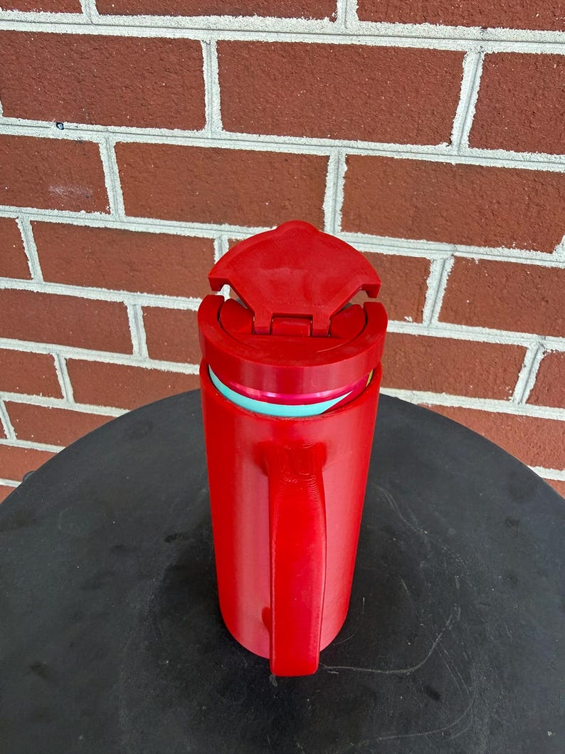 May include: A bright red insulated travel mug with a matching red handle and a hinged lid. The mug features a visible teal-colored inner rim and a cylindrical body design, providing a functional vessel for holding hot or cold beverages while on the go.