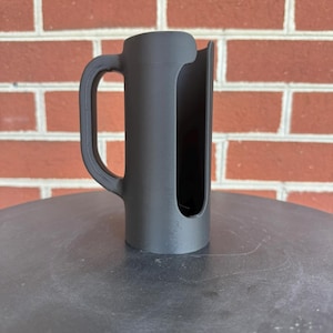 May include: A matte black cylindrical holder featuring a vertical side opening and a curved handle attached to the side. The object has a smooth, uniform finish and a hollow interior, designed with a wide cutout running along the length of the body.