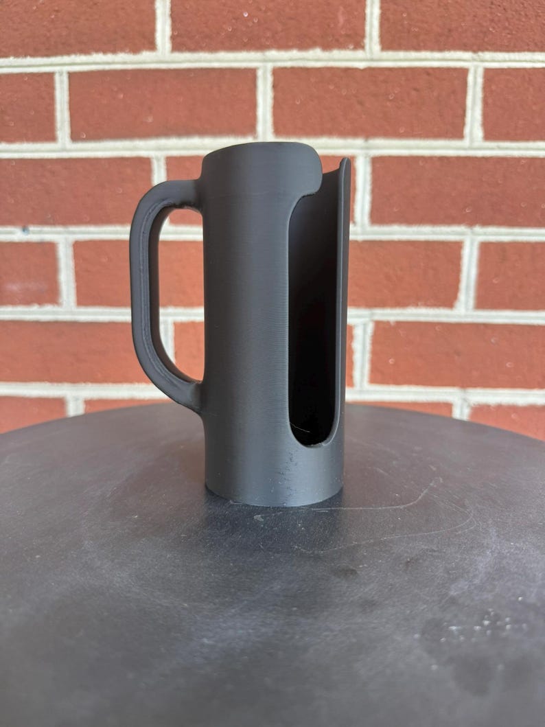 May include: A matte black cylindrical holder featuring a vertical side opening and a curved handle attached to the side. The object has a smooth, uniform finish and a hollow interior, designed with a wide cutout running along the length of the body.