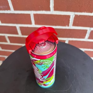 May include: A red plastic beverage can lid topper attached to the top of a slim aluminum soda can. The can features a colorful graphic design with lime illustrations and the text "Twist" printed on the side, with a silver pull tab visible.