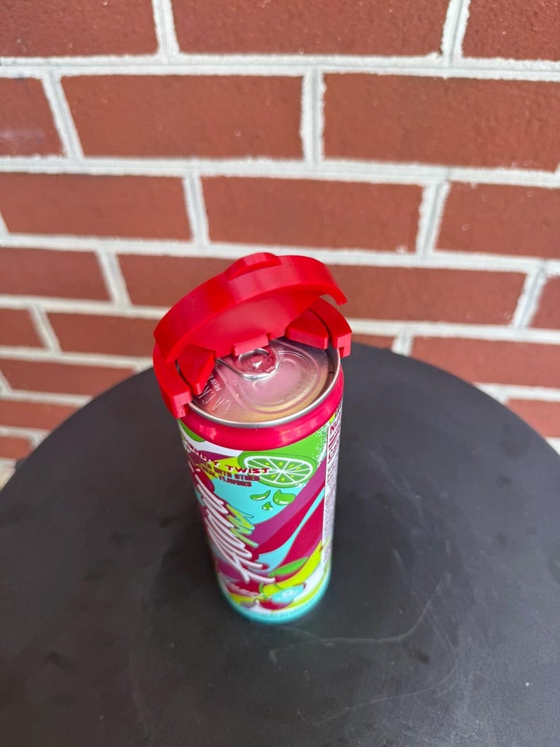 May include: A red plastic beverage can lid topper attached to the top of a slim aluminum soda can. The can features a colorful graphic design with lime illustrations and the text "Twist" printed on the side, with a silver pull tab visible.