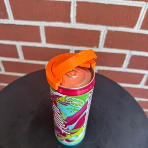 May include: A bright orange plastic beverage can handle attached to the top of a slim aluminum soda can. The can features a colorful label with green lime graphics, red and pink abstract shapes, and the visible text "Twist" in white lettering.