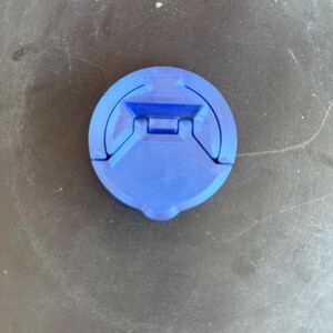 May include: A circular blue plastic cap featuring a central hinged mechanism. The cap has a smooth, matte finish with a distinct geometric design, including a recessed rectangular section and a small tab at the bottom edge for easy opening.