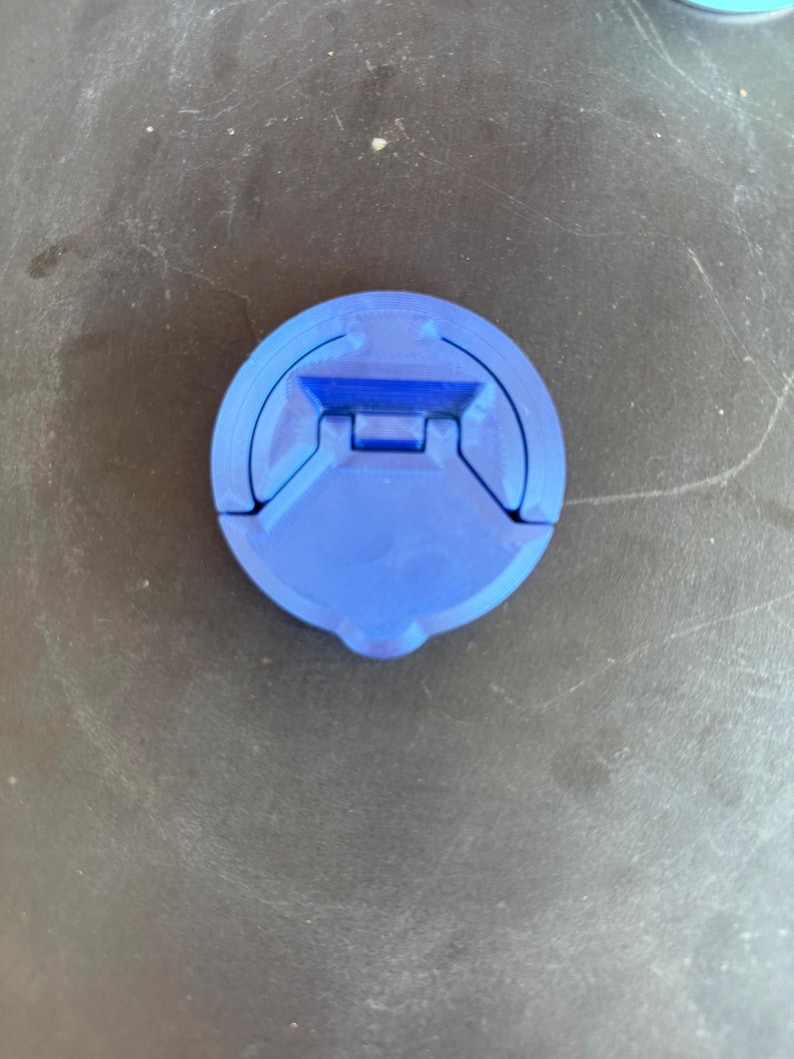 May include: A circular blue plastic cap featuring a central hinged mechanism. The cap has a smooth, matte finish with a distinct geometric design, including a recessed rectangular section and a small tab at the bottom edge for easy opening.