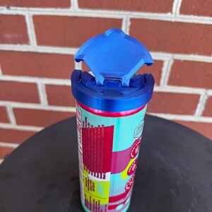 May include: A cylindrical beverage can topped with a bright blue plastic lid featuring a hinged, semi-circular flap that is currently in the open position. The can features a colorful label with red, pink, and yellow sections and visible nutritional text.