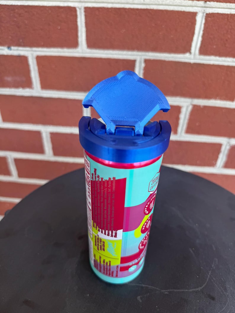 May include: A cylindrical beverage can topped with a bright blue plastic lid featuring a hinged, semi-circular flap that is currently in the open position. The can features a colorful label with red, pink, and yellow sections and visible nutritional text.