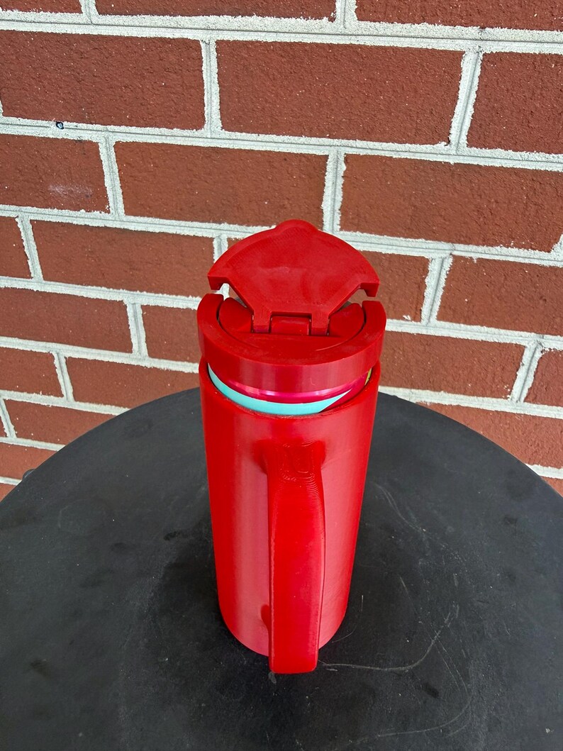 May include: A bright red insulated travel mug with a matching red handle and a hinged lid. The mug features a visible teal-colored inner rim and a cylindrical body design, providing a functional vessel for holding hot or cold beverages while on the go.