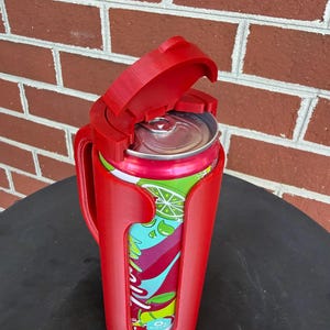 May include: A bright red plastic beverage can holder featuring a vertical handle and a hinged lid that flips open to reveal the top of a colorful soda can with green lime and red fruit graphics printed on the exterior of the aluminum container.