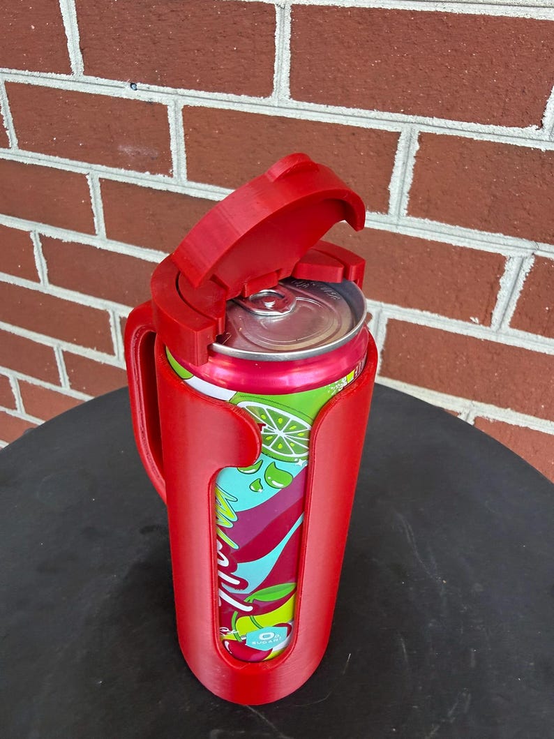 May include: A bright red plastic beverage can holder featuring a vertical handle and a hinged lid that flips open to reveal the top of a colorful soda can with green lime and red fruit graphics printed on the exterior of the aluminum container.