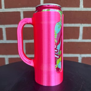 May include: A bright neon pink plastic can holder with a sturdy side handle, designed to securely fit a standard beverage can. The holder features a vertical cutout that reveals the colorful graphic design of the aluminum can inserted inside.