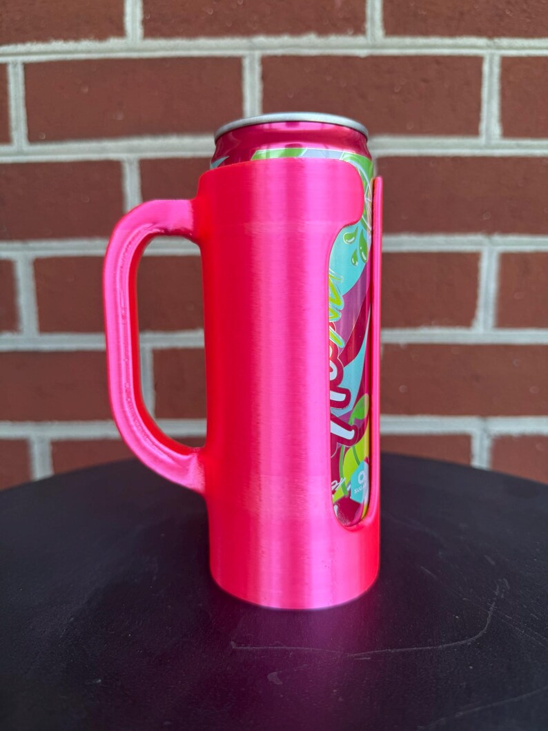 May include: A bright neon pink plastic can holder with a sturdy side handle, designed to securely fit a standard beverage can. The holder features a vertical cutout that reveals the colorful graphic design of the aluminum can inserted inside.
