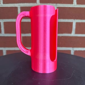 May include: A bright neon pink cylindrical holder featuring a vertical side opening and a curved handle attached to the side. The object has a smooth, uniform finish and a hollow interior, designed with a flat base for stability on a surface.