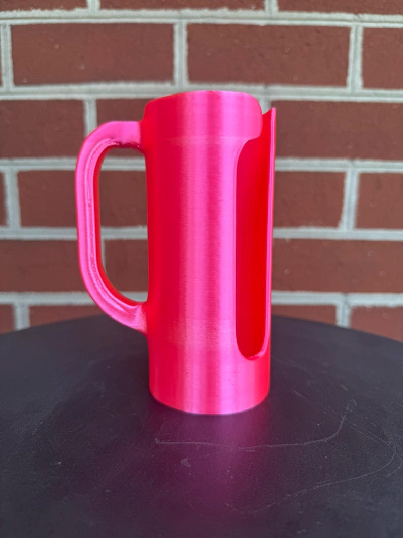 May include: A bright neon pink cylindrical holder featuring a vertical side opening and a curved handle attached to the side. The object has a smooth, uniform finish and a hollow interior, designed with a flat base for stability on a surface.