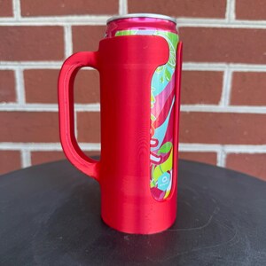 May include: A bright red plastic can holder with a sturdy side handle, designed to securely grip a slim aluminum beverage can. The holder features a vertical cutout that reveals the colorful pink, teal, and lime green graphic design on the can.
