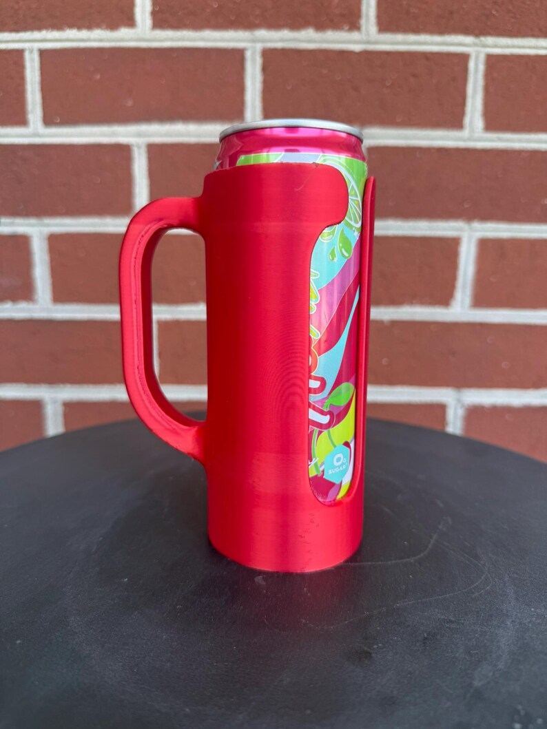 May include: A bright red plastic can holder with a sturdy side handle, designed to securely grip a slim aluminum beverage can. The holder features a vertical cutout that reveals the colorful pink, teal, and lime green graphic design on the can.