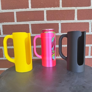 May include: Three cylindrical beverage can holders with integrated handles, arranged in a row. The left holder is bright yellow, the center holder is neon pink and contains a colorful beverage can, and the right holder is matte black.