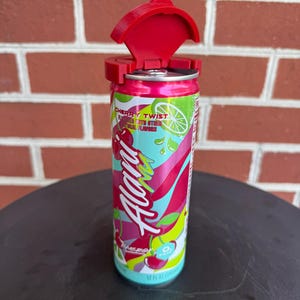 May include: A 12 fl oz Alani Nu Cherry Twist energy drink can with a bright red plastic lid attachment secured to the top. The can features a colorful graphic design with pink, green, and yellow abstract shapes, cherry illustrations, and white text.