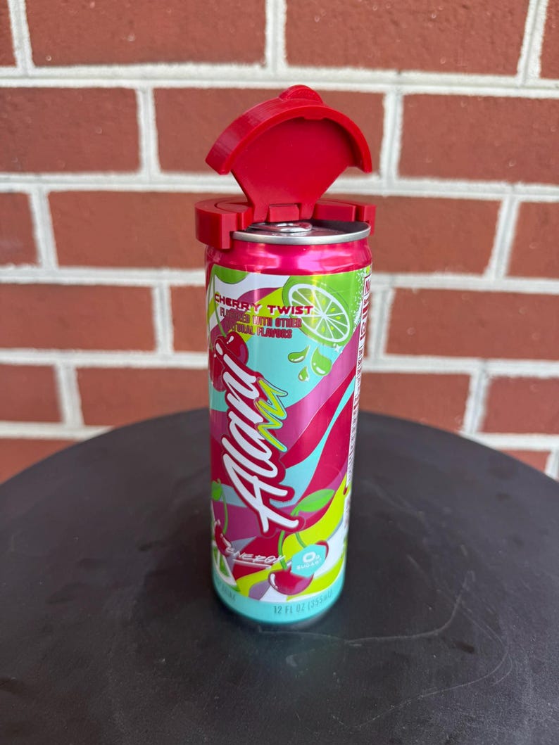 May include: A 12 fl oz Alani Nu Cherry Twist energy drink can with a bright red plastic lid attachment secured to the top. The can features a colorful graphic design with pink, green, and yellow abstract shapes, cherry illustrations, and white text.