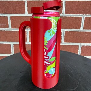 May include: A red plastic beverage can holder with a side handle and a hinged lid. The holder features a vertical cutout that reveals a colorful soda can with pink, teal, and lime green graphics, including a small white label reading 0g sugar.